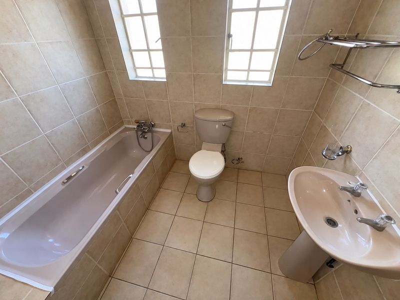 A functional bathroom featuring a bathtub, pedestal basin, and toilet with neutral floor-to-ceiling wall tiling and natural light from two windows.