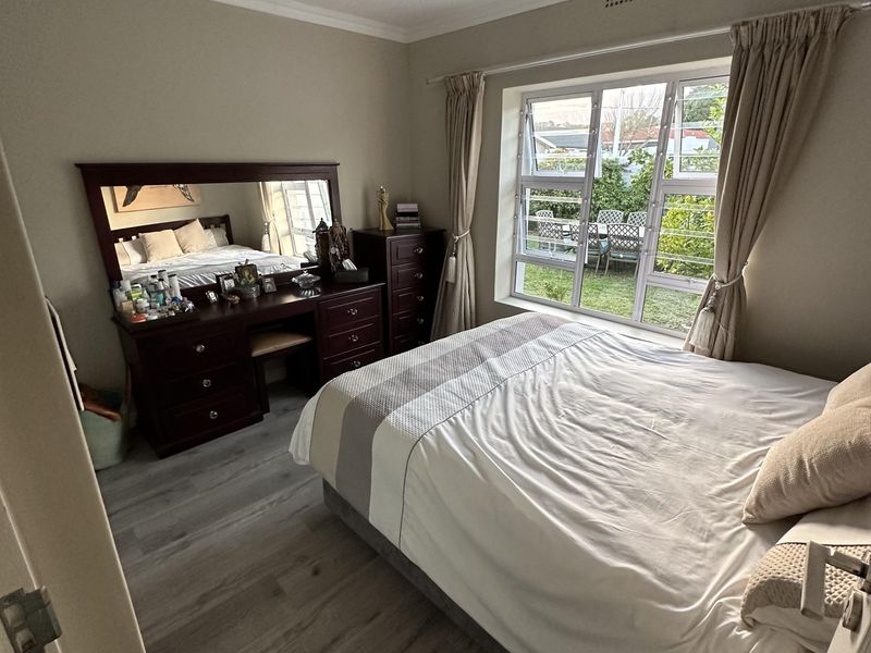 A well-maintained bedroom featuring modern laminate flooring, neutral wall tones, and a large window with security bars overlooking the garden.