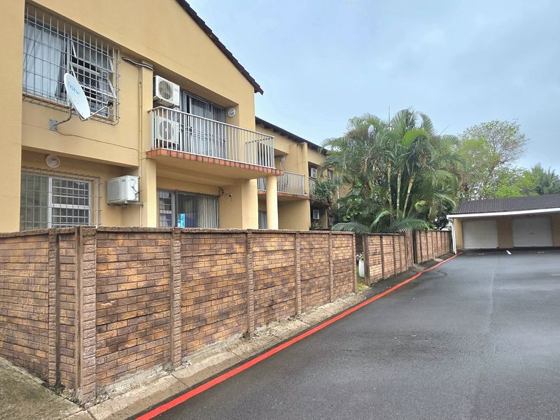 Exterior view of an apartment complex featuring brick boundary walls, balconies with security bars, and an adjacent carport area.
