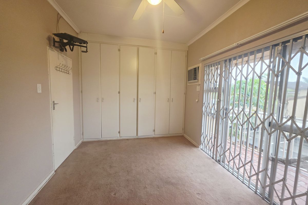 A spacious bedroom featuring built-in cupboards, a ceiling fan, and a sliding glass door with a security gate leading to an outdoor area.