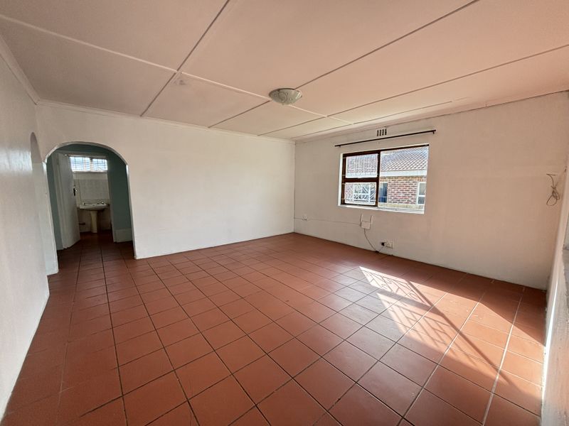 An empty living room featuring terracotta-style floor tiles, a classic archway leading to an adjacent space, and a window with security bars.
