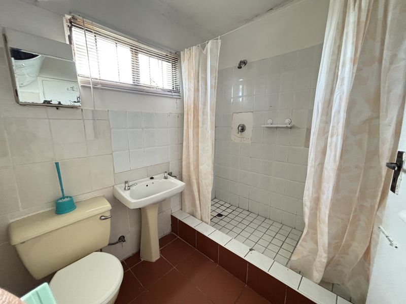 A dated bathroom featuring a pedestal basin, toilet, and a tiled shower area with a raised base, requiring renovation and cosmetic updates.