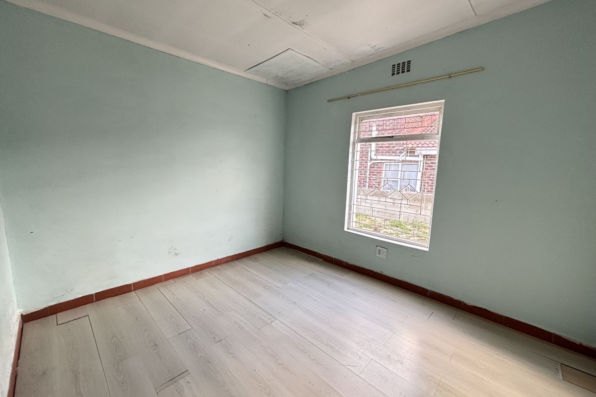 An empty bedroom featuring light-toned laminate flooring, pale blue walls, and a window fitted with security bars.