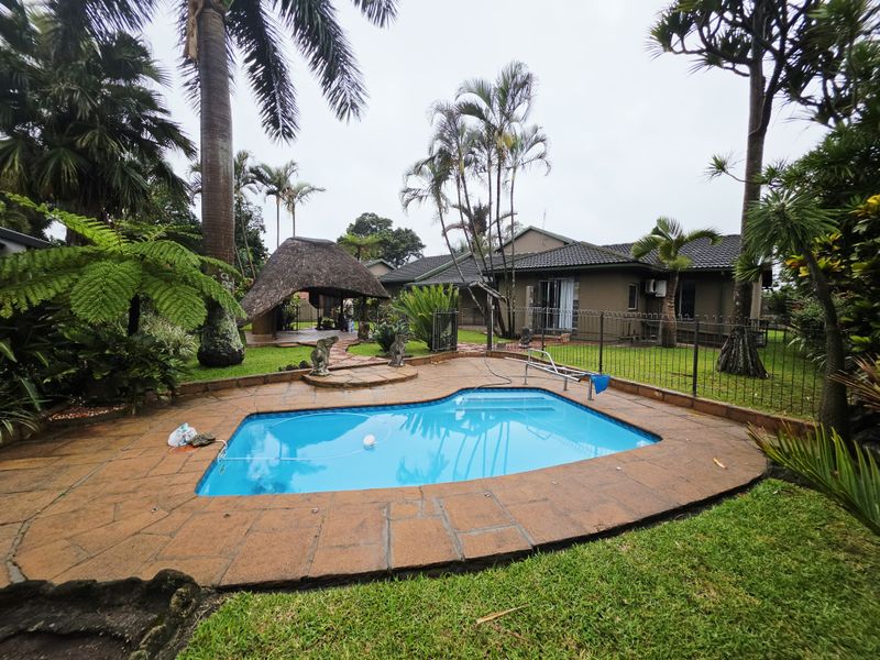 A backyard pool area featuring a swimming pool with stone paving, a thatched lapa, and established garden landscaping.
