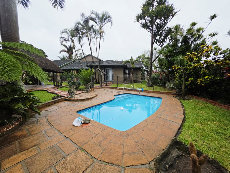 Outdoor pool area featuring a paved deck, swimming pool, and a thatched lapa set within a landscaped garden.