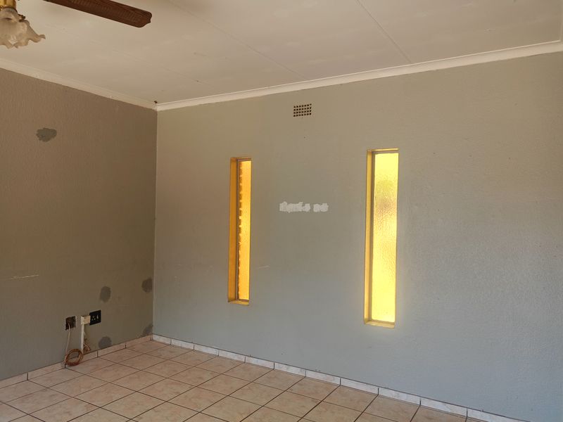 An empty living room featuring tiled flooring, vertical slit windows, and a ceiling fan, requiring cosmetic wall repairs and fresh paint.