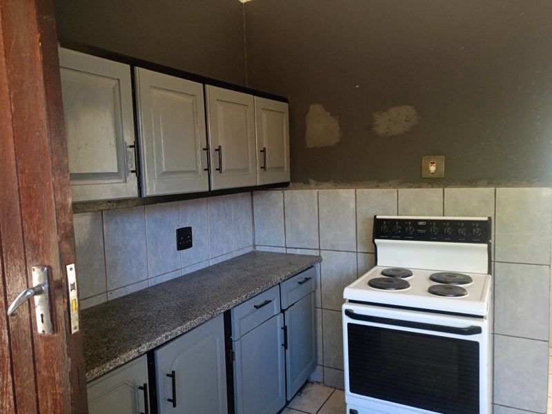 A compact kitchen featuring grey built-in cabinetry, granite countertops, tiled backsplash, and a freestanding electric stove, requiring cosmetic updates to the walls and finishes.