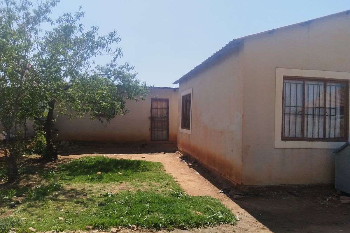 Rear view of the property showing exterior walls with security bars on windows and a metal door, set within a basic garden area.