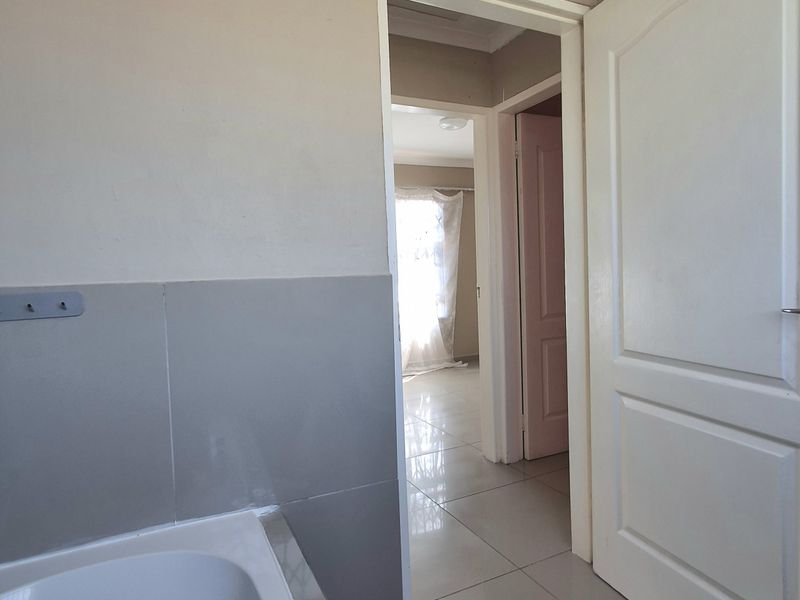 A functional bathroom featuring a built-in bathtub with tiled splashback and neutral floor tiles, leading into a hallway.