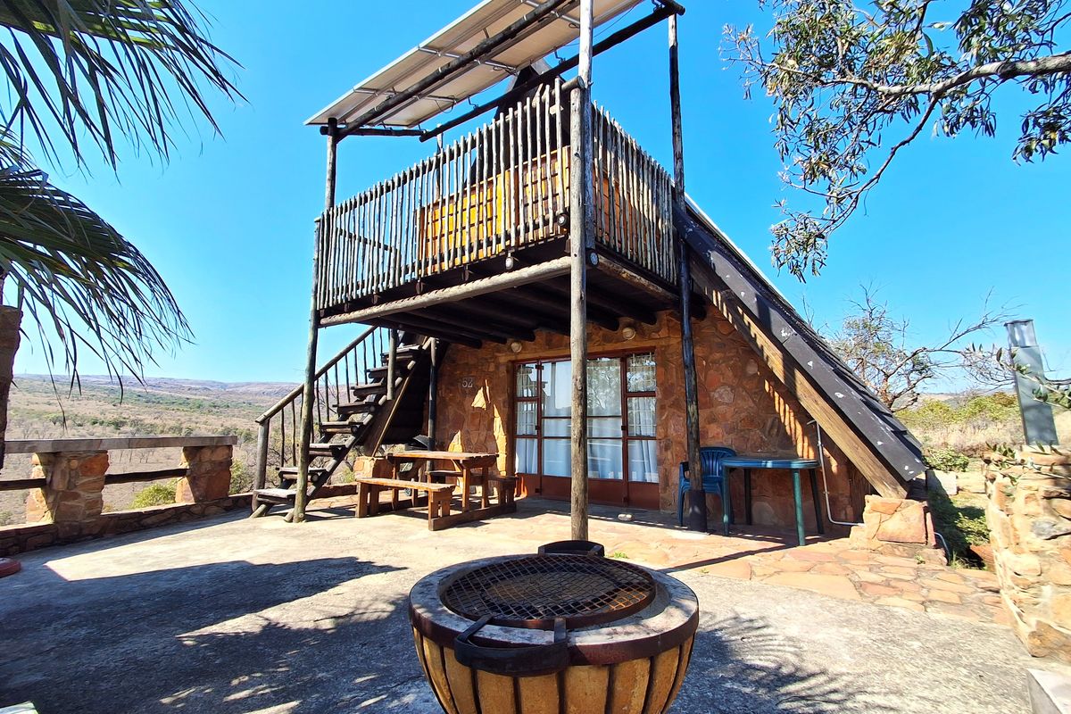 A rustic A-frame cabin featuring stone exterior walls, a raised timber balcony with solar panels, and a spacious concrete patio with a built-in braai.