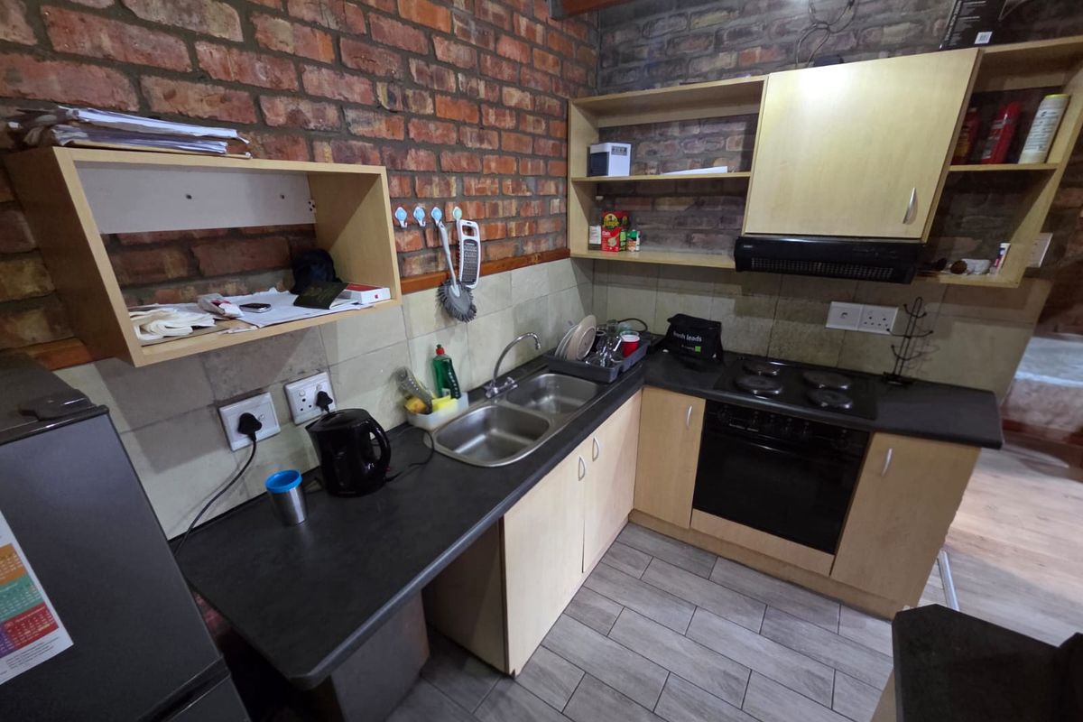 A compact kitchen featuring exposed brick walls, laminate cabinetry, a double bowl sink, and built-in cooking appliances with a tiled backsplash.