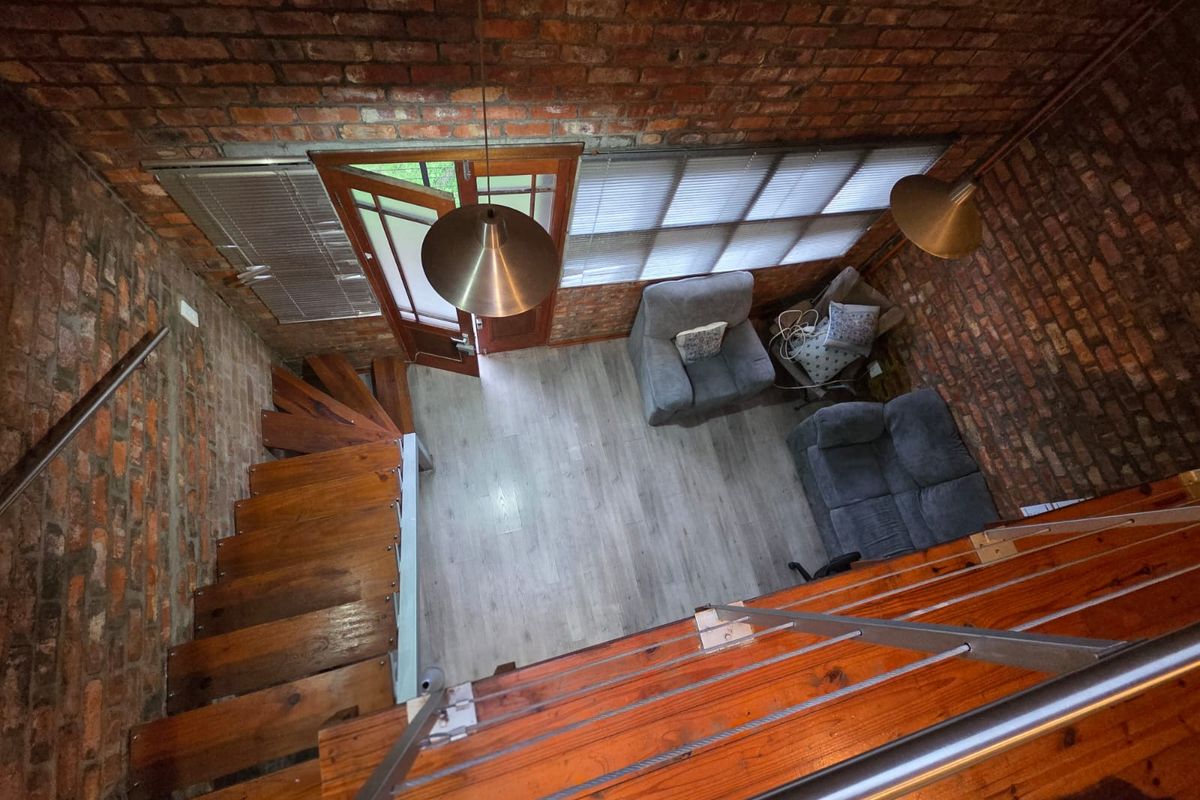 A high-angle view of a living area featuring exposed brick walls, a wooden staircase, and modern laminate flooring with natural light from the entrance.