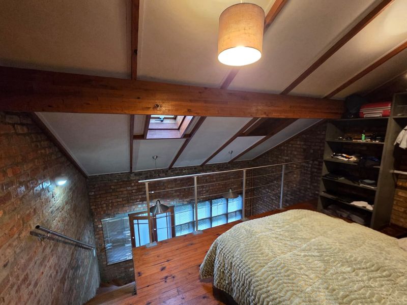 A characterful loft-style bedroom featuring exposed brick walls, timber beams, a skylight, and a mezzanine level overlooking the lower floor.