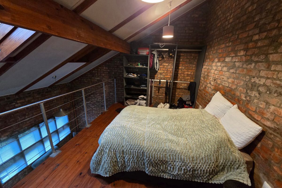 A loft-style bedroom featuring exposed brick walls, timber ceiling beams, wooden flooring, and a skylight, overlooking the lower level via a metal balustrade.