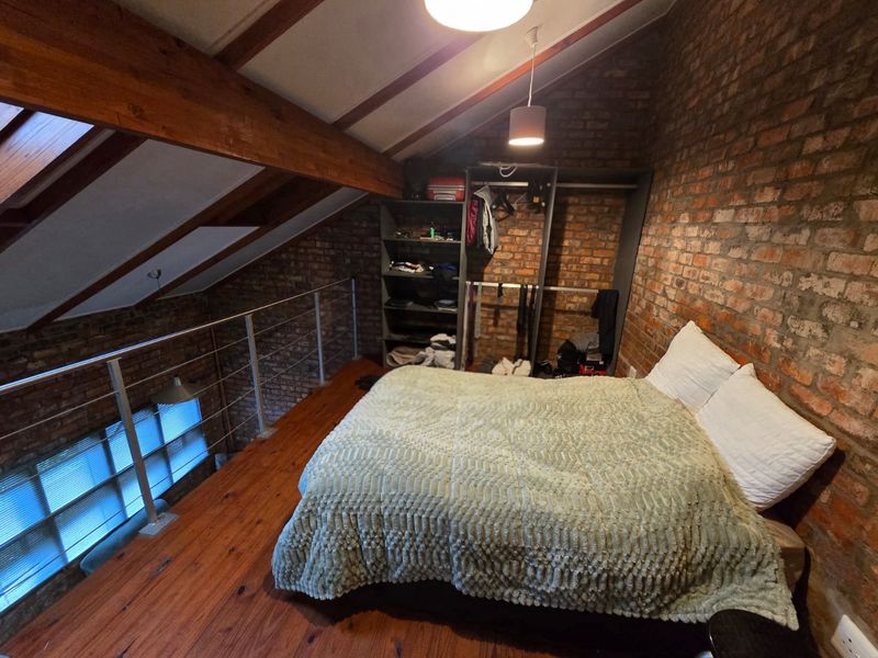 A loft-style bedroom featuring exposed brick walls, timber ceiling beams, wooden flooring, and a skylight, overlooking the lower level via a metal balustrade.