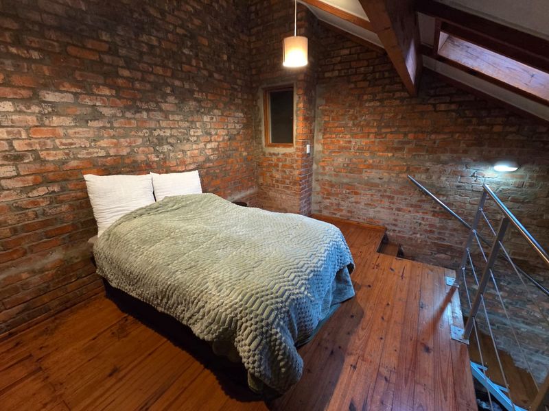 A cozy loft-style bedroom featuring exposed brick walls, warm timber flooring, exposed rafters, and a skylight, creating a unique industrial-inspired living space.