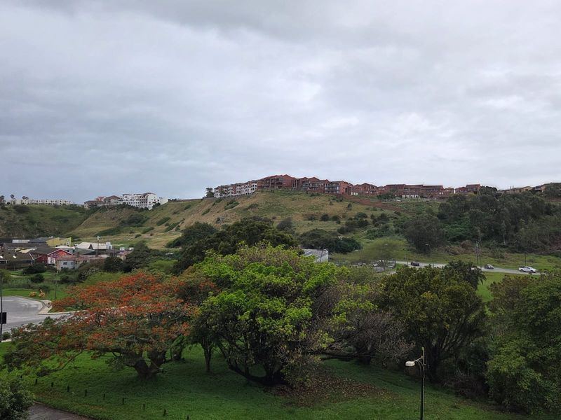 An elevated landscape view overlooking a green valley, mature trees, and distant residential apartment complexes under a cloudy sky.