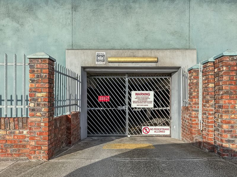 Secure gated vehicle entrance to the apartment complex parking area, featuring brick pillars and metal security fencing.