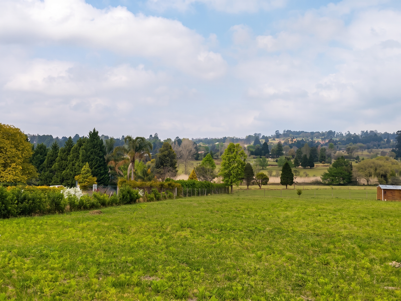 A scenic view of a large, open residential plot featuring lush green grass, established trees, and perimeter fencing under a bright sky.