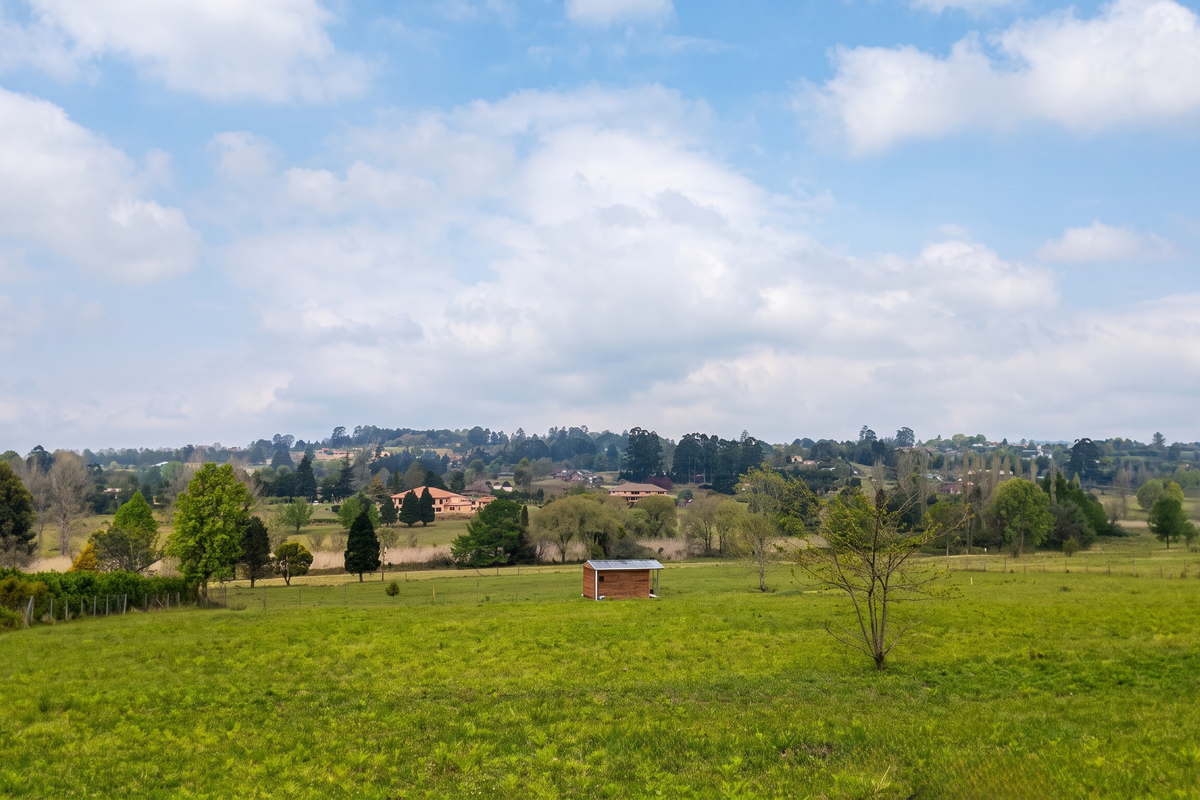 A scenic view of a large, open residential vacant land plot featuring rolling green grass, mature trees, and a small storage shed.