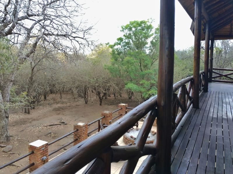 Elevated timber balcony featuring rustic wooden railings and posts, overlooking a natural bushveld landscape with a lower-level brick fire pit area.