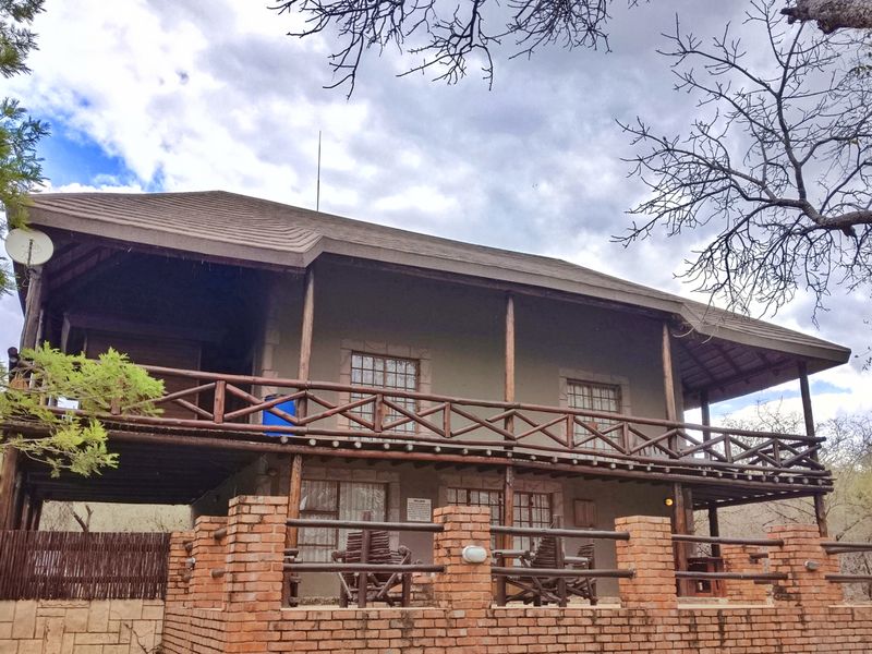 A two-story house exterior featuring a spacious wooden balcony, brick boundary wall, and rustic architectural design set within a natural bushveld environment.