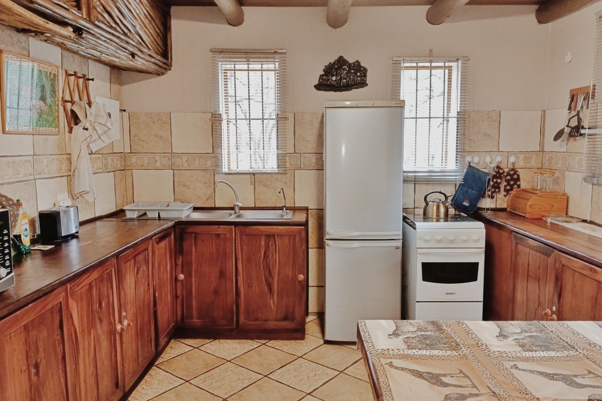 A rustic-style kitchen featuring wooden cabinetry, tiled backsplash, exposed timber ceiling beams, and a functional layout with a freestanding stove and double sink.