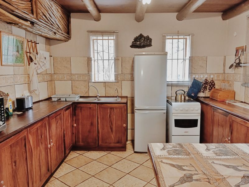 A rustic-style kitchen featuring wooden cabinetry, tiled backsplash, exposed timber ceiling beams, and a functional layout with a freestanding stove and double sink.