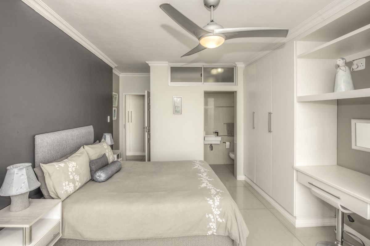 A well-maintained bedroom featuring built-in cupboards, a desk, ceiling fan, and an en-suite bathroom with tiled flooring and modern finishes.