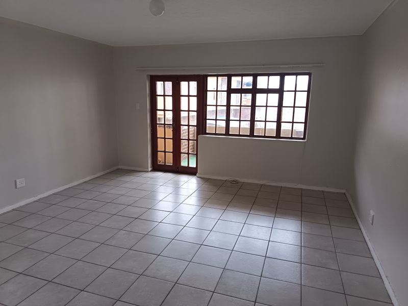 An empty, neutrally painted living room featuring tiled flooring, large multi-pane windows, and access to an outdoor area through classic french doors.