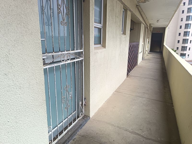 An exterior apartment corridor featuring a concrete walkway, security gate, and balustrade, showing signs of weathering and maintenance requirements.