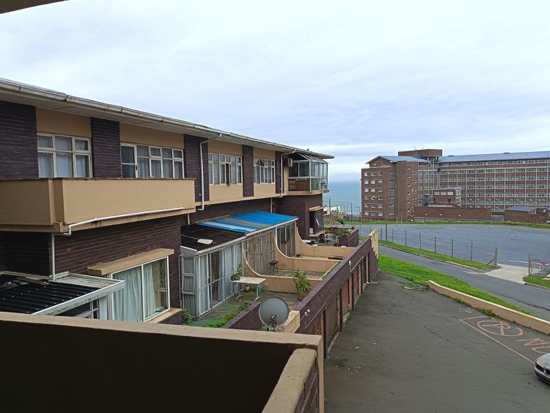 Exterior view of an apartment complex featuring brick facades, private balconies, and a paved driveway with a view towards the ocean.
