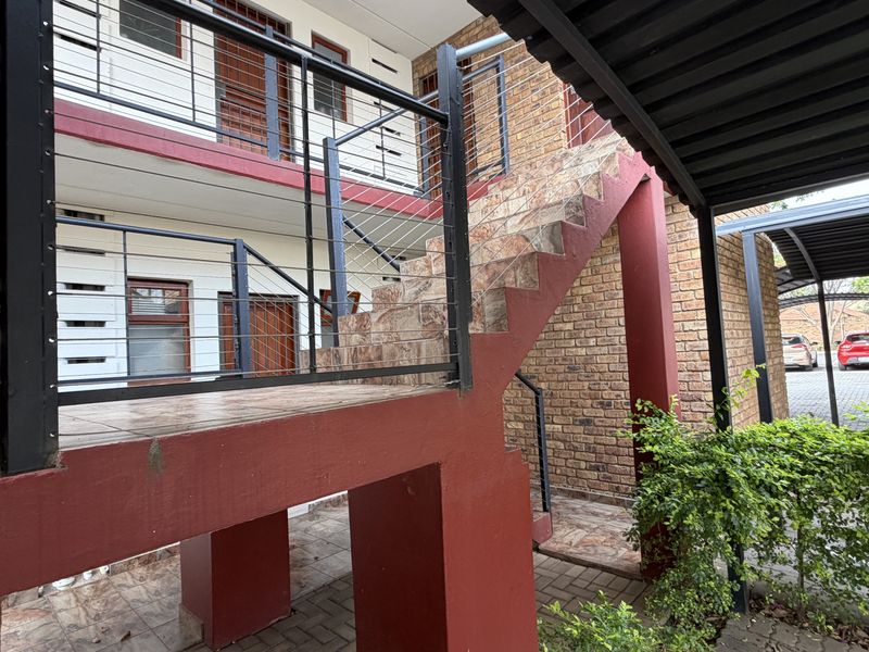 Exterior view of an apartment complex featuring a brick facade, tiled staircase with metal balustrades, and a covered carport area.