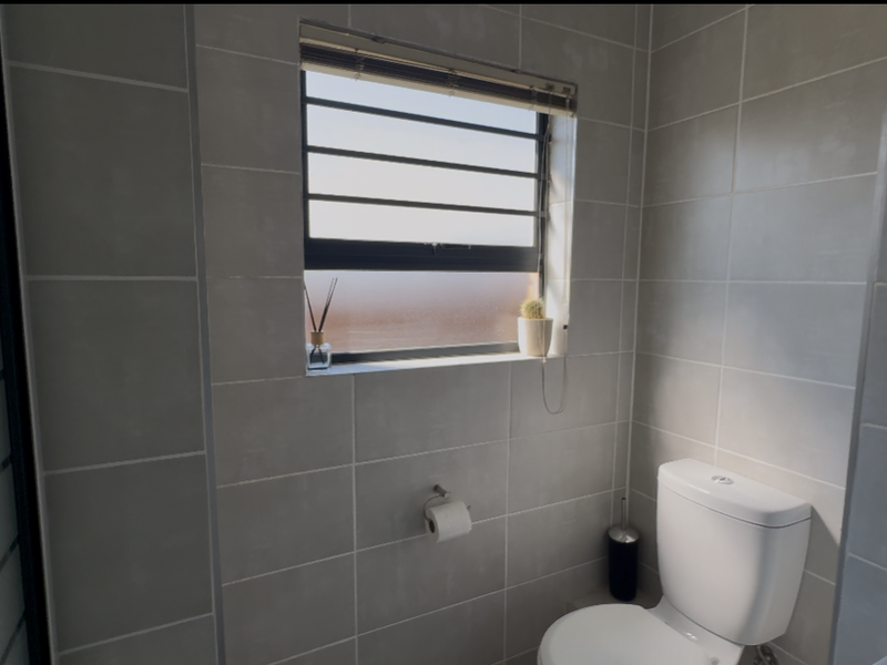 A modern bathroom featuring neutral ceramic wall tiles, a white toilet, and a frosted glass window with security bars.