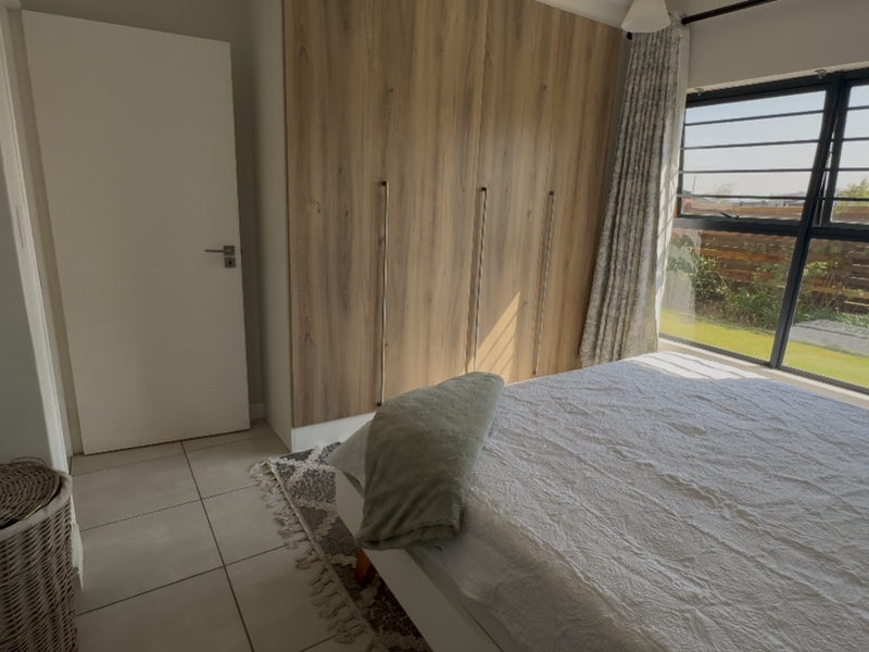 A bright bedroom featuring modern wood-finish built-in cupboards, neutral tiled flooring, and a large window providing natural light and garden views.