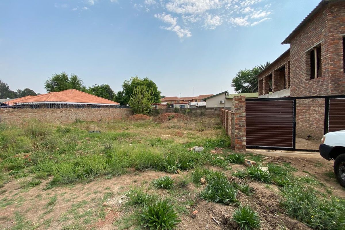 A view of a vacant residential plot featuring a brick boundary wall, metal gate, and natural vegetation.