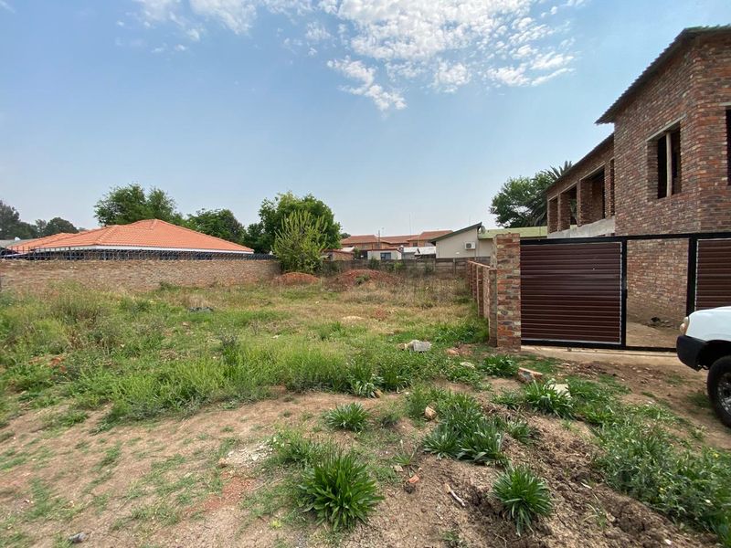 A view of a vacant residential plot featuring a brick boundary wall, metal gate, and natural vegetation.