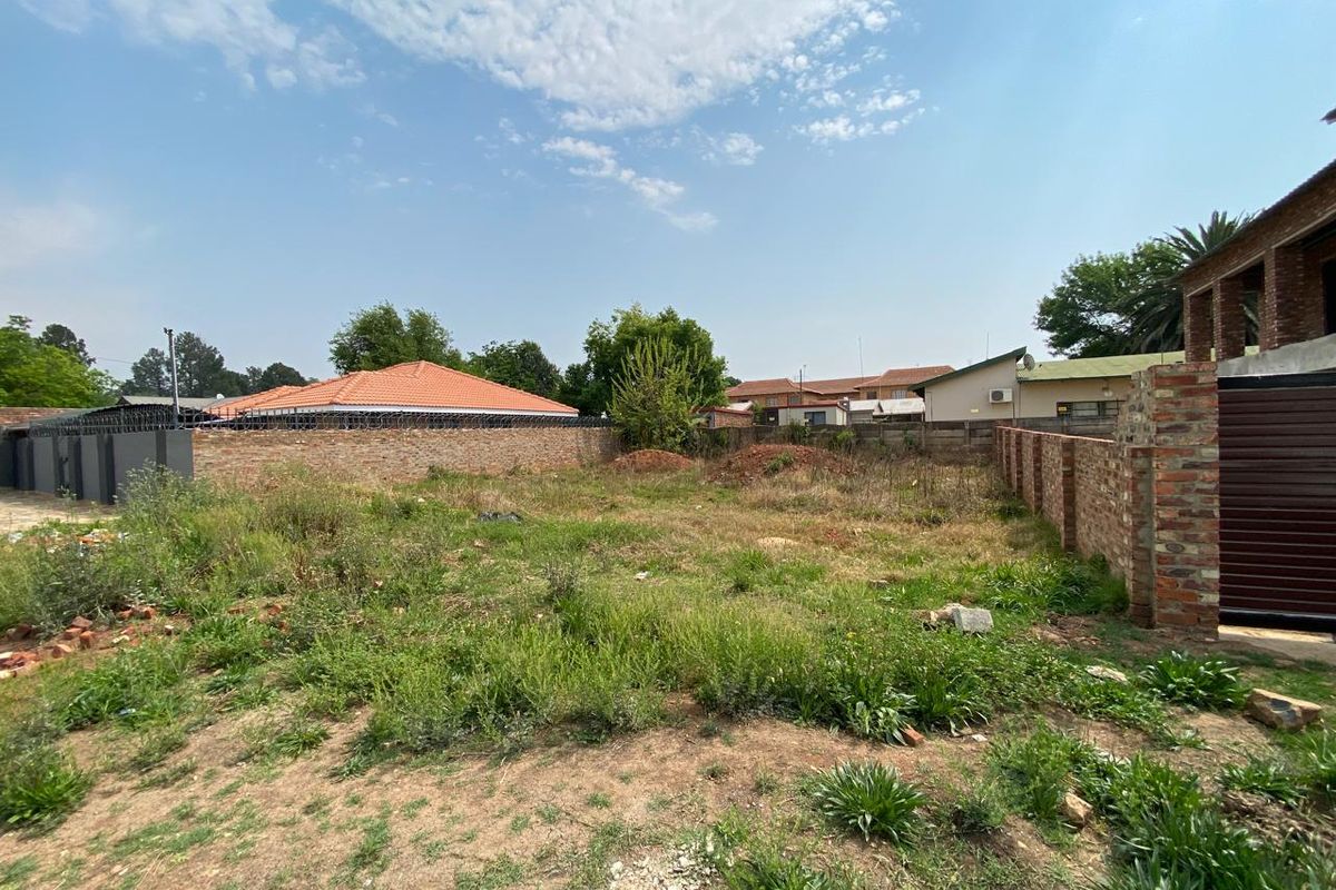 A vacant residential plot enclosed by brick perimeter walls with security spikes, featuring natural ground cover and surrounding neighborhood views.