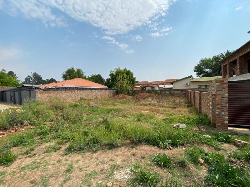 A vacant residential plot enclosed by brick perimeter walls with security spikes, featuring natural ground cover and surrounding neighborhood views.