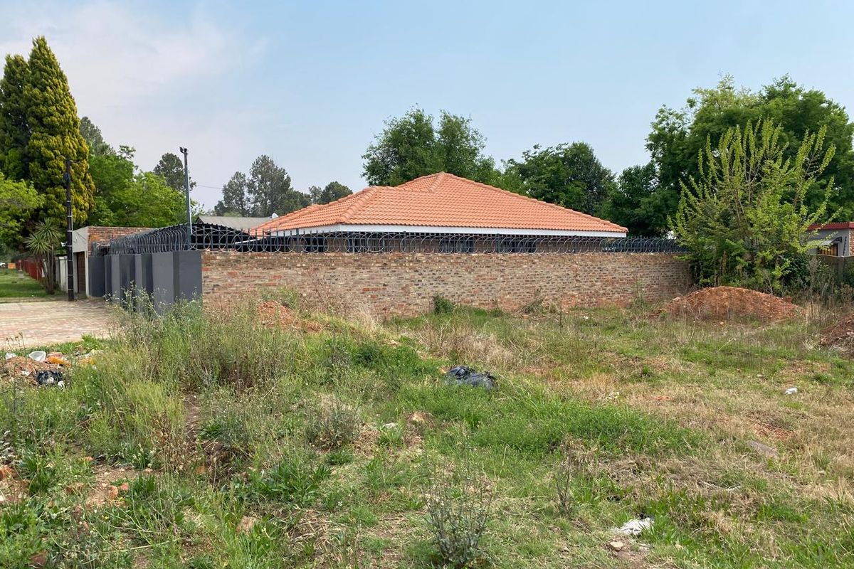 A view of a vacant residential plot featuring a brick boundary wall with security razor wire and a tiled roof structure in the background.