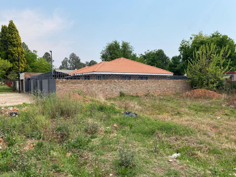 A view of a vacant residential plot featuring a brick boundary wall with security razor wire and a tiled roof structure in the background.