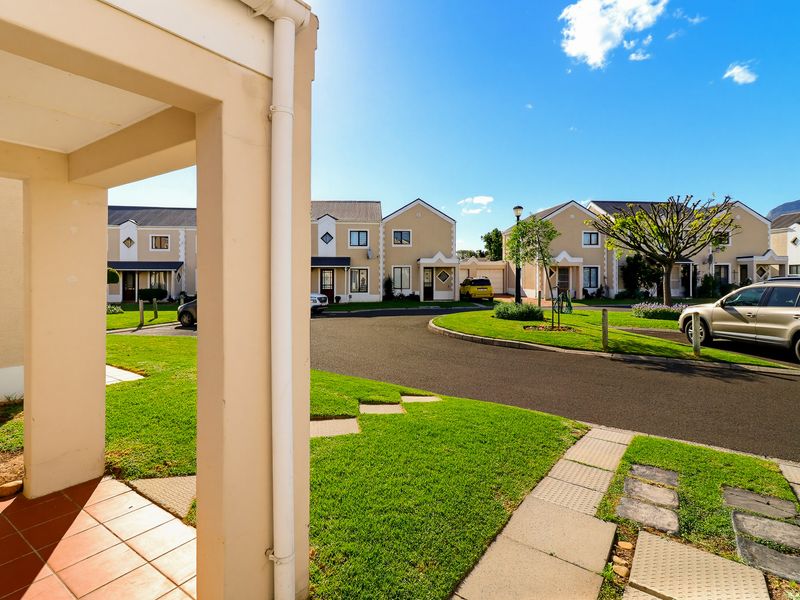 A view of the townhouse complex exterior, featuring a paved driveway, manicured lawns, and a covered tiled entrance patio.