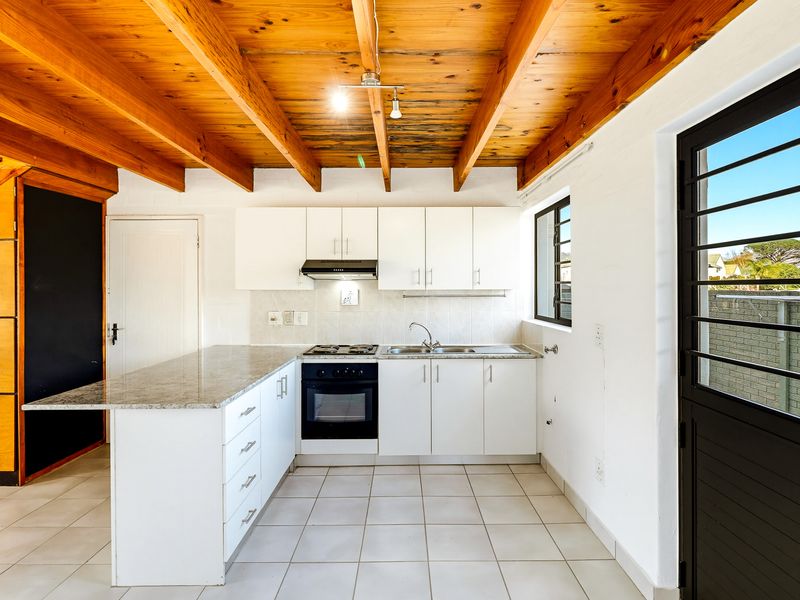 A bright kitchen featuring white cabinetry, granite countertops, built-in oven and hob, and a distinctive exposed timber beam ceiling with tiled flooring.