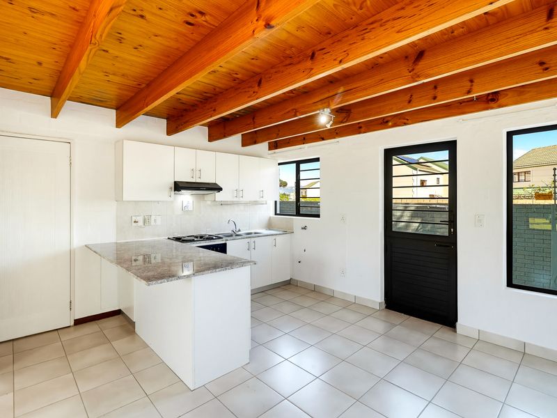 A bright, empty kitchen featuring exposed timber ceiling beams, granite countertops, built-in cabinetry, and tiled flooring with direct access to the exterior.
