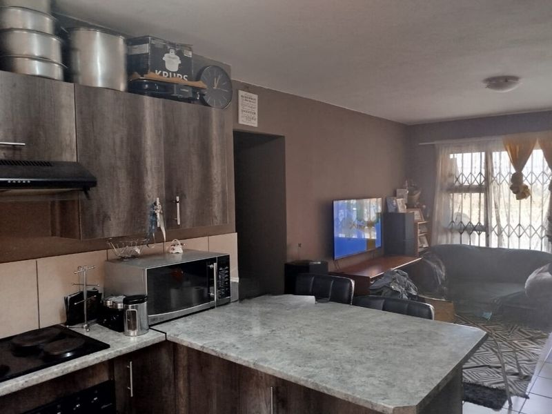 Open-plan kitchen and living area featuring modern wood-look cabinetry, stone-effect countertops, and integrated appliances, leading to a lounge with security gate access.