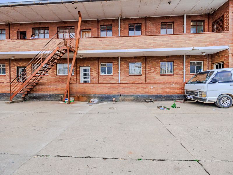 Rear exterior view of a multi-story brick apartment building featuring an external metal staircase and concrete paved ground area.