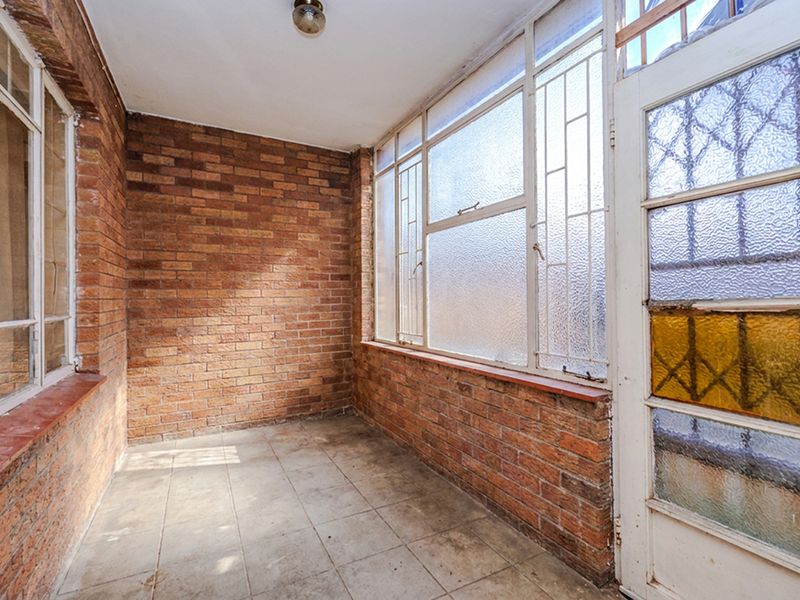 An enclosed balcony or sunroom featuring exposed brick walls, tiled flooring, and large metal-framed windows with a security door.