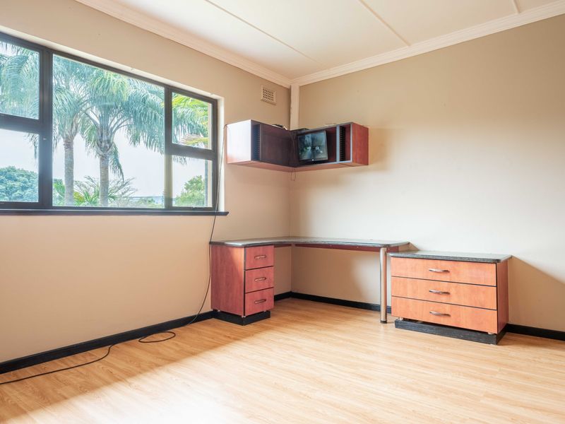 A functional home office space featuring built-in desk cabinetry, laminate flooring, and large windows providing natural light and garden views.