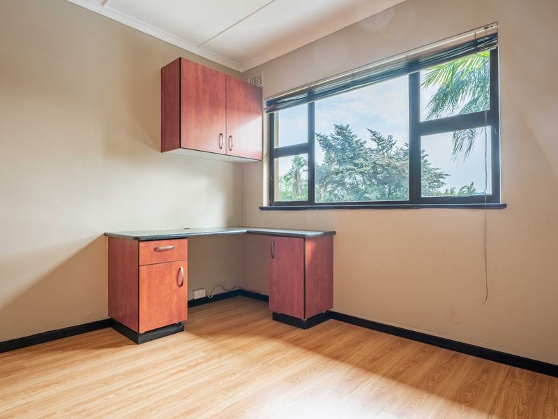 A functional home office space featuring built-in desk cabinetry, laminate flooring, and large windows providing natural light and garden views.