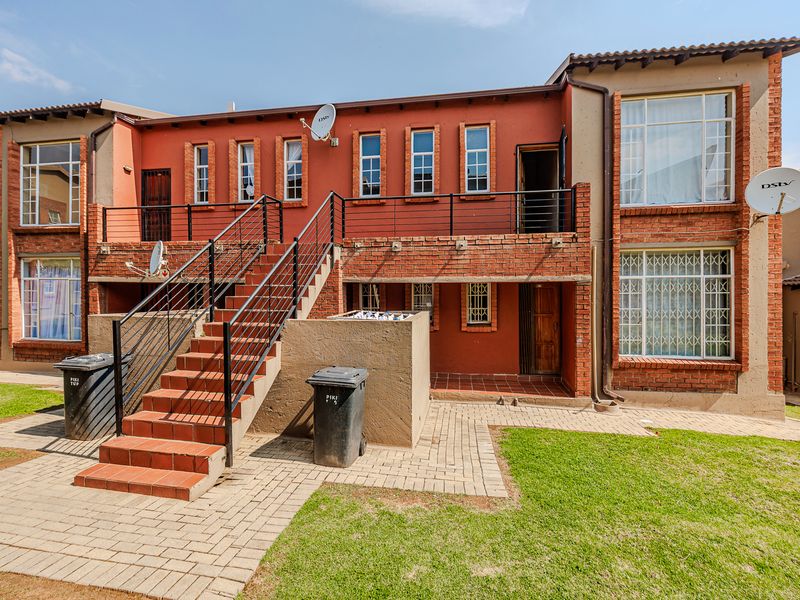 Rear view of a multi-level apartment building featuring a brick facade, external staircase with metal railings, and a paved patio area.